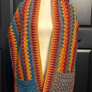 May include: A handmade, colorful crochet shawl with two pockets. The shawl features a striped pattern in shades of orange, yellow, teal, and gray. The garment is displayed on a mannequin, showcasing its design and texture. A cozy and stylish accessory.