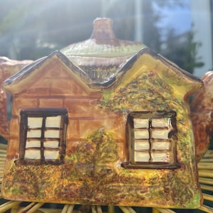 Puede incluir: Una tetera de cerámica con forma de casa, con una fachada de ladrillo marrón y dos ventanas. El tejado es de color beige y el pico y el asa son de color marrón oscuro. La tetera está sobre una superficie metálica amarilla.