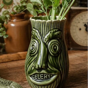 May include: A green ceramic vase shaped like a face with textured, stalk-like details and the word CELERY embossed on the mouth area. The vase has a scalloped rim and contains several stalks of fresh green celery leaves protruding from the top.
