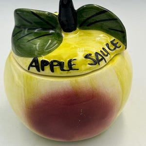 Może przedstawiać: Ceramiczny słoik w kształcie jabłka z pokrywką. Słoik jest żółty z czerwonym gradientem na dole, a pokrywka ma kształt zielonych liści. Na pokrywce czarnym kolorem napisano "APPLE SAUCE".