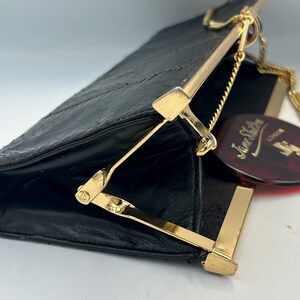May include: Black clutch bag with a gold-coloured clasp and chain. The bag has a textured surface and a rectangular shape. A red tag with the text "Jane Shilton London" is attached.