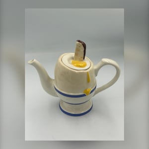 May include: A quirky ceramic teapot in an egg design. The off-white teapot has blue stripes and a handle. The lid is topped with a slice of toast and a yellow yolk. A fun and unusual addition to any kitchen.