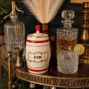 May include: A bar cart with a gin-themed setup. It features a cream-colored gin barrel with red stripes, a crystal decanter, a glass with a lime wedge, and a vintage soda siphon. The cart is adorned with decorative elements.