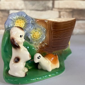 May include: A ceramic planter shaped like a boat with a dog and a turtle figurine. The boat is brown with a wood grain texture. The dog is white with brown spots and the turtle is brown and white.