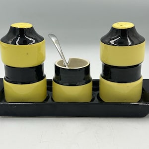 Può includere: Un set di sale e pepe nero e giallo con zuccheriera e cucchiaio abbinati, il tutto su un vassoio nero. I salini e i pepini hanno un design impilato, con fasce gialle e nere.