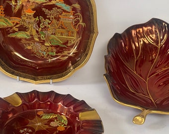 May include: Three red and gold ceramic dishes. One dish is round with a gold rim and a painted scene of a Chinese village. Another dish is shaped like a leaf with a gold rim. The third dish is round with a gold rim and a painted spider web.