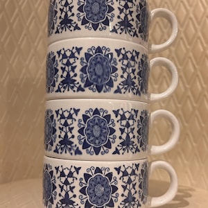 May include: A stack of four white ceramic mugs with a blue floral pattern. Each mug has a rounded shape, a handle, and a repeating design of stylized flowers and leaves. The mugs are stacked neatly, showcasing the intricate blue and white design.
