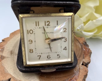 May include: A vintage travel clock in a black leather case. The clock face is white with black numerals and hands, and a gold-colored frame. The clock reads "TEMPORA" and "7 JEWELS GERMANY". The case is open, revealing the clock.