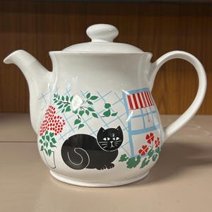 May include: White ceramic teapot featuring a black cat design, red and green floral patterns, and a blue and white grid. The teapot includes a lid and a curved handle.