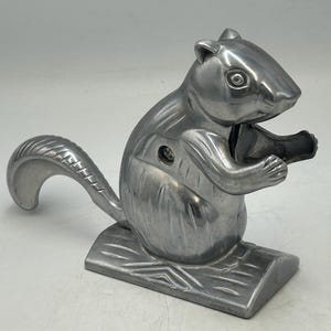 May include: A silver-coloured metal nutcracker shaped like a squirrel sitting on a textured base. The squirrel has a curved tail, defined facial features, and front paws positioned as if holding an object. The metal has a smooth, polished finish.