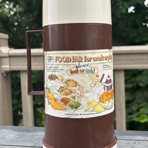 Può includere: Un thermos vintage marrone e crema con manico. L'etichetta recita "FOOD JAR for work or play" e "Serve hot or cold". Il thermos presenta illustrazioni di vari alimenti. Prodotto da Thermos Ltd., Brentwood, Essex, Inghilterra.