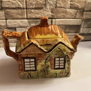 Può includere: Una teiera in ceramica marrone e gialla a forma di cottage con tetto di paglia. La teiera ha due finestre e un camino marrone.