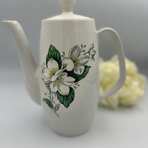 May include: Off-white ceramic teapot with a floral pattern. The teapot has a white base, adorned with white flowers, green leaves, and grey details. The lid and handle are white, with a gold rim on the lid. A classic tea accessory.