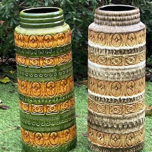May include: Two tall, cylindrical ceramic vases with textured, repeating patterns. One vase is green and gold, the other is brown and gold. Both have a wide, open top and are displayed on green grass.