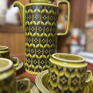 Puede incluir: Un juego de café vintage verde y negro que incluye una cafetera, tazas y platillos. La cafetera tiene una forma alta y cilíndrica con tapa y asa. Las tazas y los platillos tienen un diseño a juego. El juego tiene una estética retro.