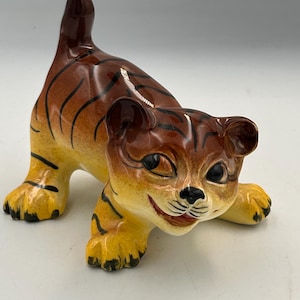 May include: A ceramic tiger figurine in a playful pose. The tiger has a brown and yellow color scheme with black stripes. The figurine has a smiling expression and is on all fours.