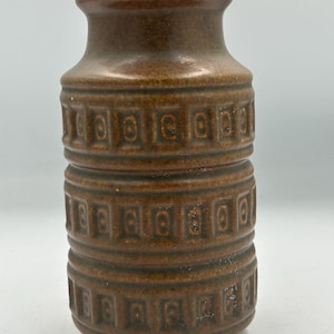 Może przedstawiać: Brązowy ceramiczny wazon o cylindrycznym kształcie i wąskiej szyi. Wazon posiada powtarzający się geometryczny wzór wokół korpusu. Kolor jest ciepły, ziemisty brąz, a powierzchnia ma lekko teksturowany wygląd. Ten dekoracyjny wazon nadaje się do dekoracji wnętrz.
