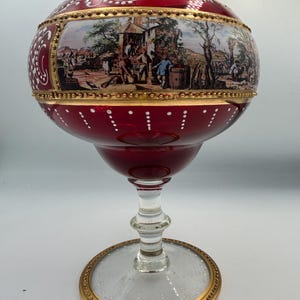 May include: A ruby red glass goblet with a wide bowl and a clear, circular base. The bowl features a painted scene framed by gold trim. White dots and decorative swirls adorn the goblet's exterior. The base and rim are trimmed with gold.