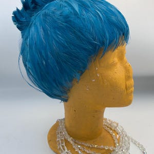 Puede incluir: Un tocado de plumas azul vibrante, probablemente un fascinador o sombrero, se exhibe sobre una cabeza de maniquí amarilla. Las plumas están densamente empaquetadas, creando una apariencia texturizada. Un collar de cuentas transparentes de múltiples hilos adorna el cuello del maniquí.