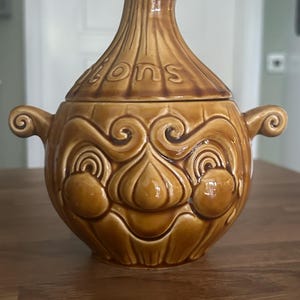 Può includere: Un contenitore a forma di cipolla in ceramica marrone con coperchio. Il contenitore ha un design a forma di viso con manici su ogni lato. La parola "Onions" è in rilievo sul coperchio. Il contenitore è appoggiato su una superficie di legno.