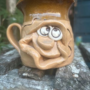 Könnte beinhalten: Eine handgefertigte Keramiktasse mit einem skurrilen Gesichtsdesign. Die Tasse hat einen beigefarbenen Sockel mit einem dunkleren braunen Rand und Henkel. Das Gesicht hat große, ausdrucksstarke Augen und einen lächelnden Mund, was der Keramik einen spielerischen Touch verleiht.