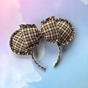 May include: A headband featuring two round, padded ears and a central bow, all covered in a brown, white, and black plaid fabric. The ears and the bow are finished with a ruffled edge, and the headband frame is thin and flexible.