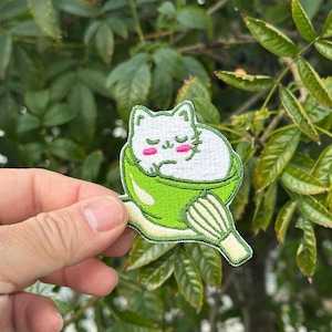 May include: Embroidered patch featuring a white cat sleeping in a green matcha bowl with a whisk. The cat has pink cheeks and closed eyes. The patch is surrounded by a green border and is approximately 2.5 inches wide.