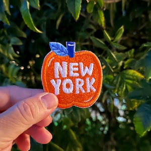 May include: An embroidered patch shaped like an orange apple with a blue stem and leaf. The text NEW YORK is stitched in white capital letters across the center of the apple. The patch features a white border and a textured, thread-based finish.