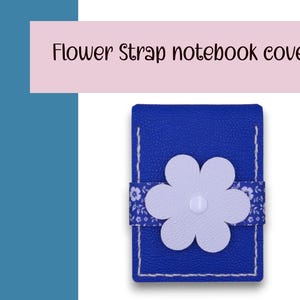 May include: A blue notebook cover with a white flower strap. The cover has white stitching and a floral patterned strap. The text "Flower Strap notebook cover" is displayed above the notebook.