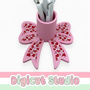 May include: A pink bow-shaped desk organizer with a cylindrical holder for pens or tools. The bow's wings are decorated with a red and white heart pattern. The organizer is on a white background.
