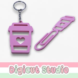 May include: A pink keychain and paperclip set. The keychain is shaped like a coffee cup with a heart cutout, attached to a silver ring. The paperclip is also pink, with a coffee cup design and a heart cutout. The background is white.