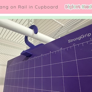 May include: A purple cutting mat with a grid pattern and measurement markings, hanging from a white closet rod. The mat has the text "StrongGrip" and numbers indicating inches. A blue hanger is used to suspend the mat.
