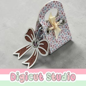 May include: A miniature gift bag with a floral pattern, cream bow, and a brown bow-shaped charm. The bag features a handle and metal eyelets. The charm is connected with a metal clasp. The text "Digicut Studio" is at the bottom.