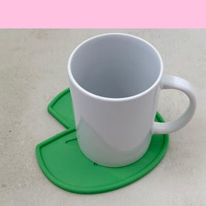 May include: A white ceramic mug with a curved handle sits on a bright green, leaf-shaped coaster. The coaster has a rounded edge and a cutout design. The background is a light gray surface with a pink upper border.