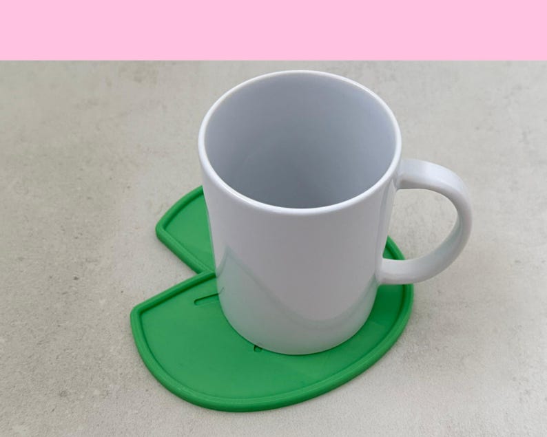 May include: A white ceramic mug with a curved handle sits on a bright green, leaf-shaped coaster. The coaster has a rounded edge and a cutout design. The background is a light gray surface with a pink upper border.