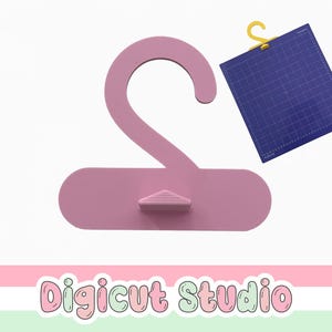 May include: A light pink clothing hanger with a small triangular support. A blue cutting mat with a yellow hook is to the right. The text "Digicut Studio" is at the bottom in a colorful font.
