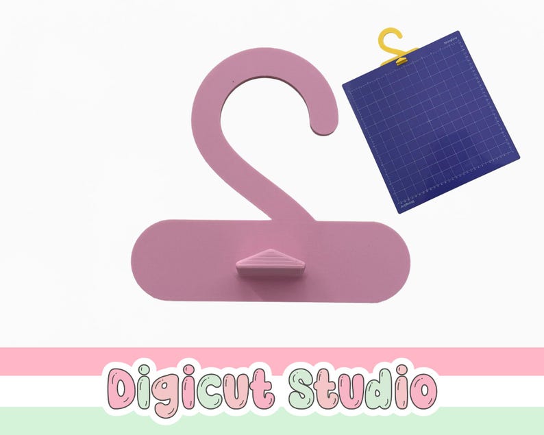 May include: A light pink clothing hanger with a small triangular support. A blue cutting mat with a yellow hook is to the right. The text "Digicut Studio" is at the bottom in a colorful font.