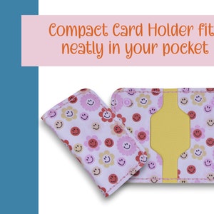 May include: A pink card holder with a yellow interior and a pattern of pink and yellow flowers and smiley faces. The text "Compact Card Holder fits neatly in your pocket" is printed above the card holder.
