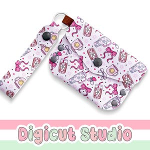 May include: A white card holder with a repeating pattern of pink bows, strawberries, and beverage tumblers with flowers. The accessory features a matching wrist strap, a metal ring, and black snap buttons. The text Digicut Studio is visible.