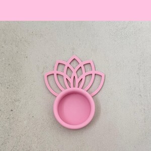 May include: A pink lotus flower-shaped coaster. The coaster has a circular base with a floral design above it. The coaster is made of a solid pink material. The background is a light gray color.