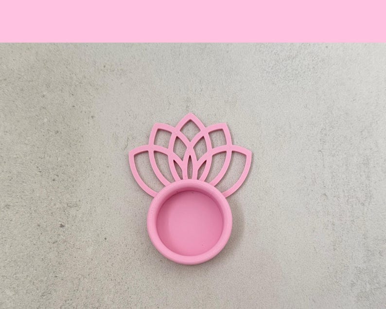 May include: A pink lotus flower-shaped coaster. The coaster has a circular base with a floral design above it. The coaster is made of a solid pink material. The background is a light gray color.