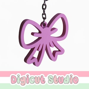 May include: A lavender-colored bow-shaped keychain hanging from a silver chain. The bow has a cutout design and is suspended against a white background. The text "Digicut Studio" is at the bottom.