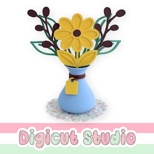 May include: A blue vase featuring a yellow sunflower and brown stems. The vase has a small yellow tag and rests on a white doily. The image also includes the text "Digicut Studio" in pink and green.