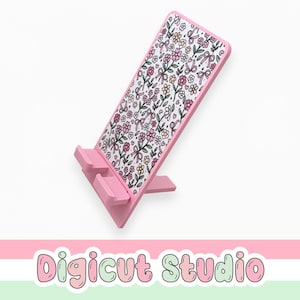 May include: A pink phone stand with a floral pattern. The stand is angled and features a white background with a repeating pattern of pink, yellow, and green flowers and bows. The base has a support for the phone.