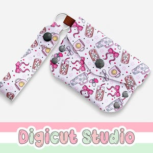 May include: A white key holder and wristlet set featuring a pattern of pink bows, coffee cups, and drinks. The key holder has a brown leather strap and black snap closures. The wristlet has a matching pattern and a black snap closure. The text "Digicut Studio" is at the bottom.