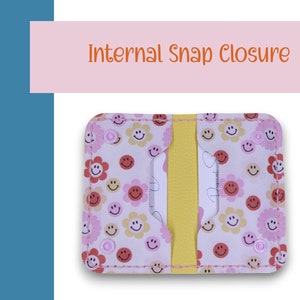 May include: A white leather card holder with a pink and yellow smiley face pattern. The card holder has a yellow elastic band and a snap closure.