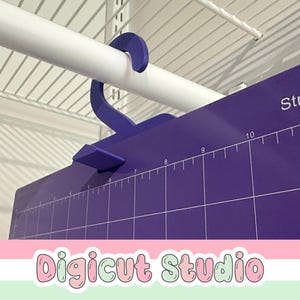 May include: A purple cutting mat with white measurement markings and the word "Strong" printed on the top right. A purple hook is attached to the mat, hanging from a white rod. The text "Digicut Studio" is at the bottom.