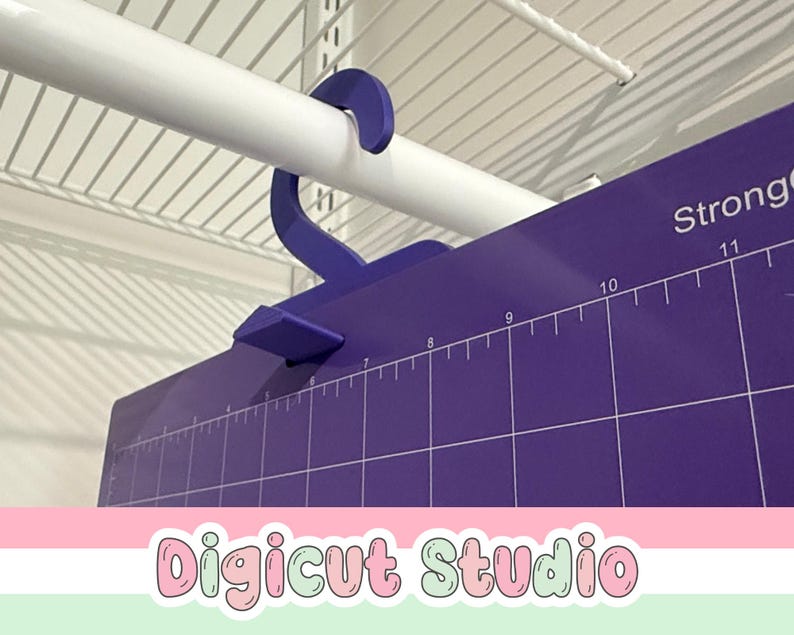 May include: A purple cutting mat with white measurement markings and the word "Strong" printed on the top right. A purple hook is attached to the mat, hanging from a white rod. The text "Digicut Studio" is at the bottom.