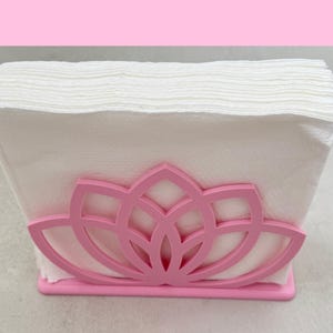 May include: A pink napkin holder with a lotus flower design, holding a stack of white napkins. The holder is made of a solid material and has a rectangular base.