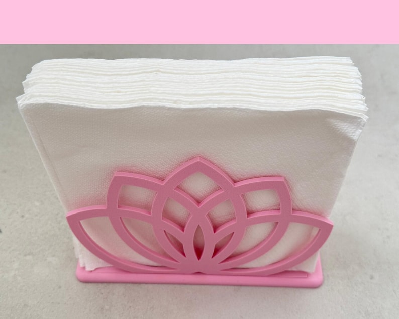 May include: A pink napkin holder with a lotus flower design, holding a stack of white napkins. The holder is made of a solid material and has a rectangular base.